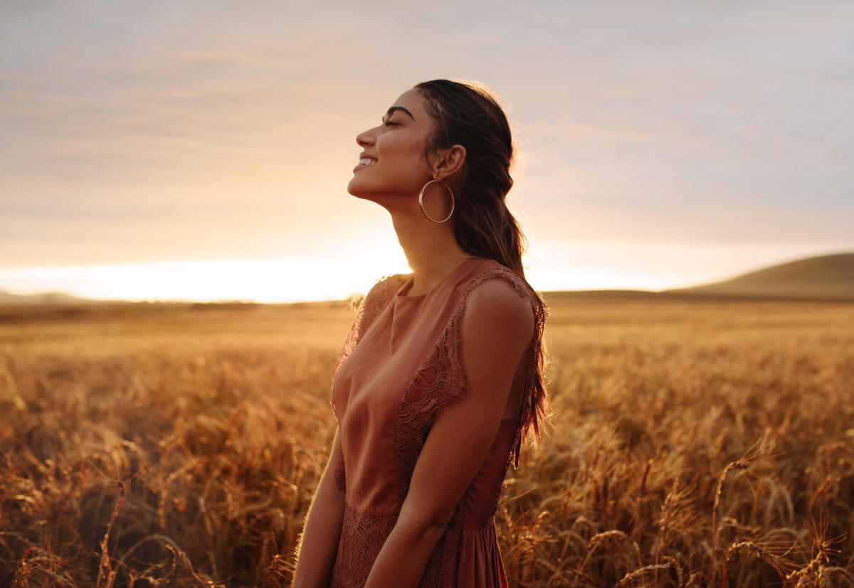 A woman standing in a field of wheat during a sunset is smiling and calming enjoying the moment with her eyes closed.