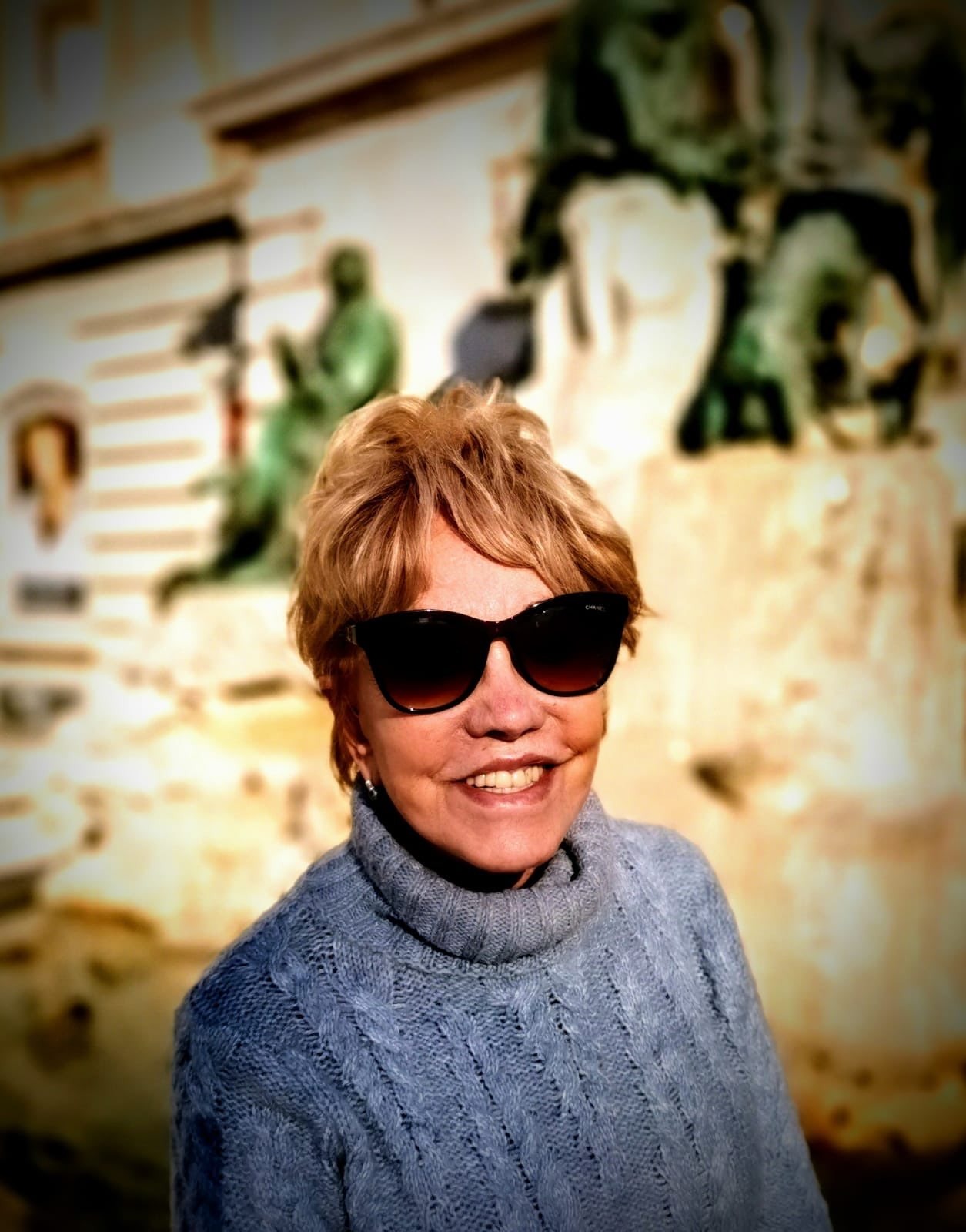 A photograph of Eliana, a woman standing in the sun whilst wearing sun glasses and smiling.