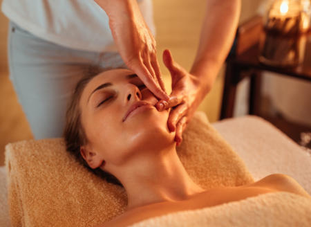 A relaxed woman wearing a towel is having her cheek massaged whilst her eyes are closed.