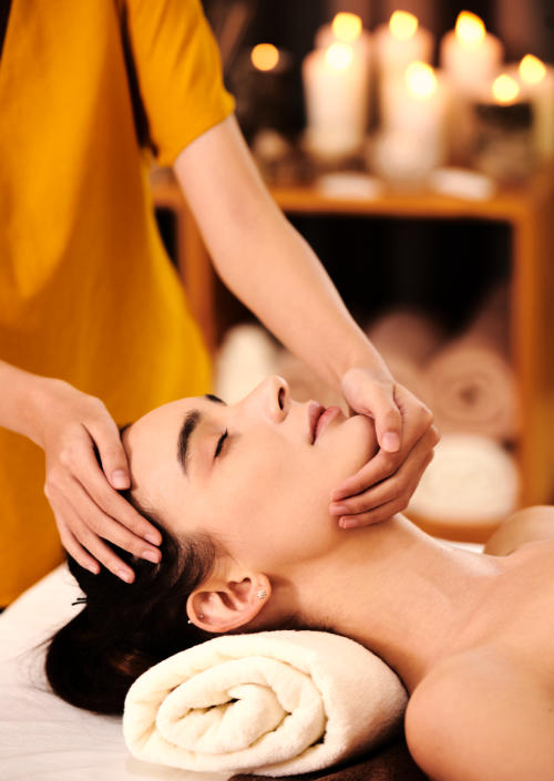 A relaxed woman with closed eyes getting her face massaged with her neck resting on a
				pillow.