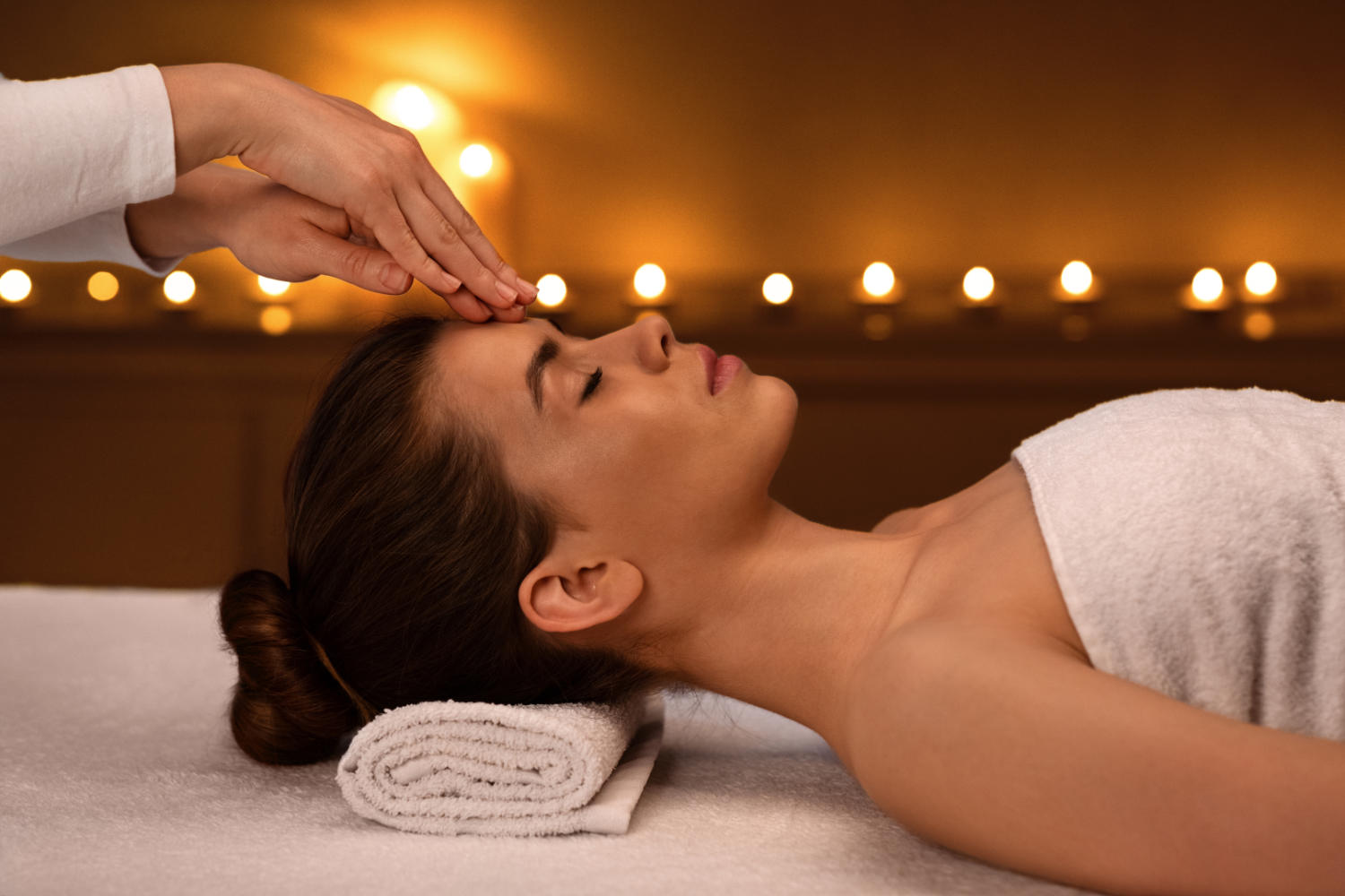 A relaxed woman with closed eyes is getting her forehead massaged with candles in the
				background.